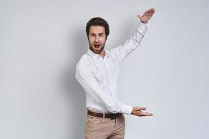 Surprised young man in white shirt looking at camera and gesturing while standing against gray background photo
