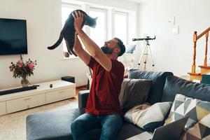 Charming young man in casual clothing bonding with domestic cat and smiling while spending time indoors photo