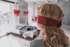 Something is going to surprise her. Rear view of young woman standing blindfold in the bedroom full of balloons and rose petals photo