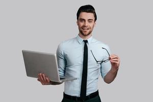 Always ready to help.  Young handsome man in white shirt and tie holding laptop and eyeglasses while standing against grey background photo