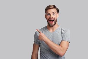 Charming young man pointing copy space and smiling while standing against grey background photo