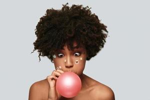 Funky mood. Surprised young African woman blowing the balloon while standing against grey background photo