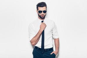 Confident handsome. Handsome young man in white shirt adjusting his necktie and looking at camera while standing against white background photo