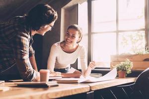 Discussing new project. Cheerful young man and woman in casual wear pointing blueprint and communicating while both standing near wooden desk in creative office photo