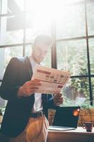 Man reading newspaper. Low angle view of confident young man reading fresh newspaper while leaning at the window sill in office or cafe photo