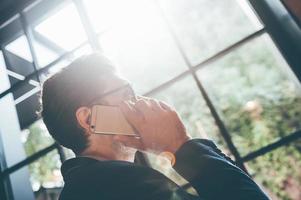 Man on the phone. Low angle view of young man talking on the mobile phone and looking away while standing in front of the big window indoors photo