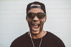 disfrutando de su música favorita. apuesto joven africano con auriculares mirando a la cámara y manteniendo la boca abierta mientras está de pie frente a la pared de piedra al aire libre foto