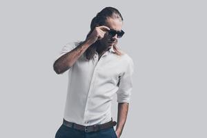 elegante y guapo. un joven apuesto con camisa blanca ajustando sus gafas de sol mientras se enfrenta a un fondo gris foto