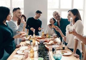 buena fiesta. grupo de jóvenes con ropa informal comiendo pizza y sonriendo mientras cenan en el interior foto