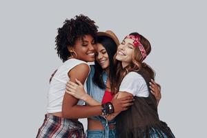 tres atractivas y elegantes mujeres jóvenes abrazándose y riéndose mientras se enfrentan a un fondo gris foto