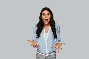 Real shock Surprised young woman gesturing and looking at camera while standing against grey background photo