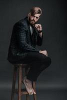 Stylish and handsome. Full length of attractive young man looking at camera and holding hand on chin while sitting on chair against grey background photo