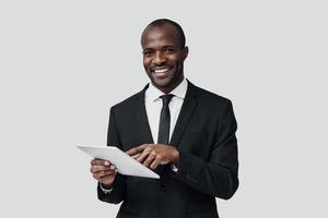 Charming young African man in formalwear working using digital tablet while standing against grey background photo