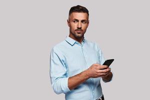 Searching for an answer. Good looking young man in smart casual wear using his smart phone and smiling while standing against grey background photo