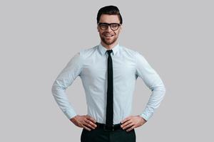 Young and successful. Good looking young man in eyeglasses keeping hands on hip and looking at camera while standing against grey background photo