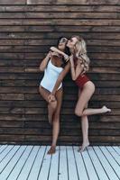 Happy and carefree. Full length of two attractive young women in swimwear laughing and bonding while standing against the wooden wall outdoors photo