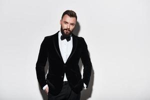 Perfect style. Handsome young man in full suit keeping hands in pockets and looking at camera while standing against grey background photo