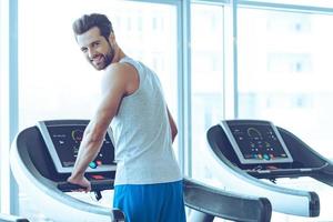 Handsome on treadmill. Rear view of young handsome man in sportswear standing on treadmill in front of window at gym and looking at camera with smile photo