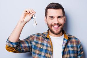 Happy house owner. Smiling young man holding keys and looking at camera while standing against grey background photo
