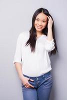 Shy beauty. Beautiful young Asian woman looking at camera and holding hand in hair while standing against grey background photo