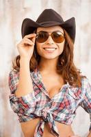 chica de campo. hermosa joven vaquera ajustando sus gafas y sonriendo a la cámara mientras está de pie contra el fondo de madera foto