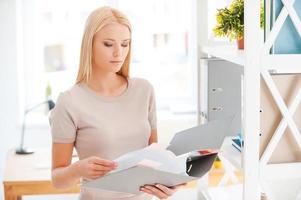 Looking for the right document. Beautiful young woman examining documents while standing near the shelf in office photo