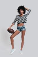 Playful woman. Full length studio shot of attractive young African woman in casual wear keeping hand in hair and carrying a ball while standing against grey background photo