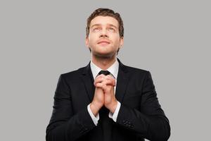 Businessman praying. Thoughtful young man in formalwear holding hands clasped and fingers crossed while standing against grey background photo