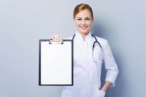 Copy space on her clipboard. Confident female doctor in white uniform holding clipboard and looking at camera while standing against grey background photo