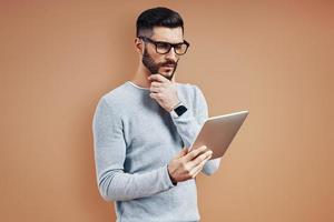 Thoughtful young man in smart casual wear keeping hand on chin and using digital tablet while standing against brown background photo
