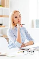 New day new decisions. Pensive young beautiful woman holding hand on chin and looking thoughtful while sitting at her working place photo