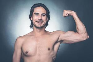 fuerte y guapo. apuesto joven sin camisa mostrando su bíceps perfecto y mirando a la cámara con una sonrisa mientras se enfrenta a un fondo gris foto