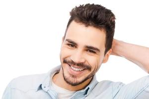 sonrisa sincera apuesto joven tocando su cabeza y sonriendo mientras está de pie contra el fondo blanco foto