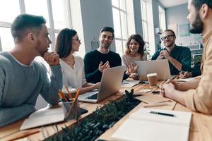 confiado e inteligente. grupo de jóvenes modernos con ropa informal inteligente discutiendo algo y sonriendo mientras trabajan en la oficina creativa foto