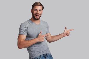 Take a look over here. Charming young man pointing copy space and smiling while standing against grey background photo