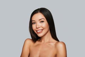 Shiny beauty. Beautiful young Asian woman looking at camera and smiling while standing against grey background
