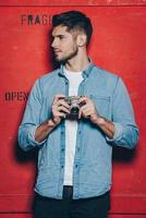 Handsome photographer. Handsome young man in sunglasses keeping arms crossed and looking at camera while standing against red background photo