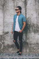 Confident in his style. Full length of handsome young man in cap and sunglasses standing against the concrete wall photo