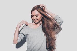 alegre belleza. atractiva mujer joven cogida de la mano en el cabello y sonriendo mientras está de pie contra el fondo gris foto