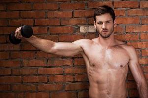 Showing his strength. Serious young muscular man training with dumbbells while standing against brick wall photo