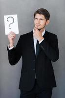 He has big question. Thoughtful young businessman holding note pad with question mark on it and looking away while standing against grey background photo