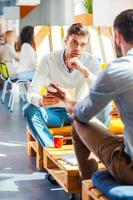 Business conversation. Two confident young men in smart casual wear discussing something while sitting in office with their colleagues working in the background photo