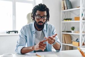 Confident young African man in eyewear telling something and looking at camera while working indoors photo