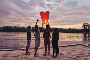 Nothing is better than old friends. Full length of young people in casual wear preparing sky lantern while standing on the pier photo