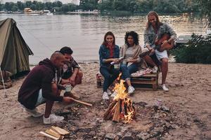 Getting away from it all... Group of young people in casual wear smiling while enjoying beach party near the campfire photo