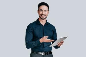 Always ready to help. Handsome young man in shirt pointing on his digital tablet and smiling while standing against grey background photo