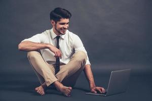 Staying online. Full length of confident young handsome man using laptop with smile while sitting on the floor against grey background photo