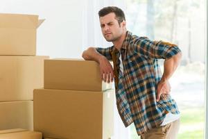 Feeling pain in back. Frustrated young man holding hand on his back and expressing negativity while leaning at the cardboard box photo