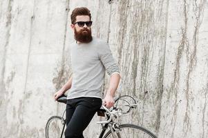 Street style. Pensive young bearded man leaning at his bicycle and looking away while standing against the concrete wall photo