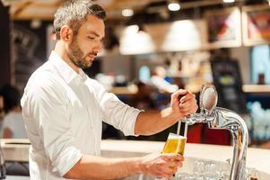 Pouring beer for client. Side view of young bartender pouring beer while standing at the bar counter photo